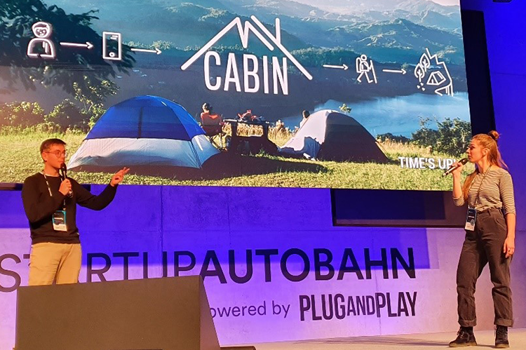 Finn Wilkesmann (links im Bild) und Sophia Hummler (rechts im Bild) stehen auf einer Bühne beim Pitch im Rahmen des Academic Seed Accelerator Program (ASAP) von Start-Up BW im Februar. Auf der Leinwand hinter ihnen ist das „MyCabin“-Logo auf einem Foto der Alpen abgebildet.