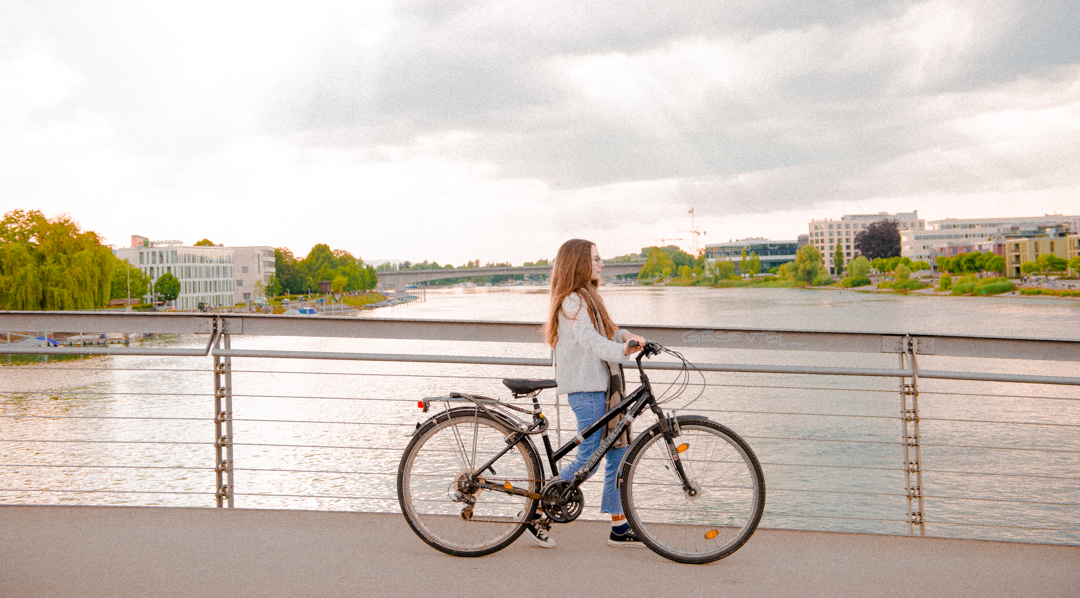 Eine junge Frau läuft über die Fahrradbrücke in Konstanz und schiebt ein Fahrrad. Im Hintergrund ist die HTWG zu sehen.