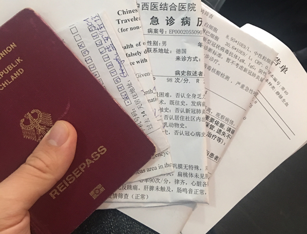 In einer Hand liegen verschiedene Dokumente aufgefächert. Unter einem deutschen Reisepass sind das weiße Papiere mit chinesischen Schriftzeichen.