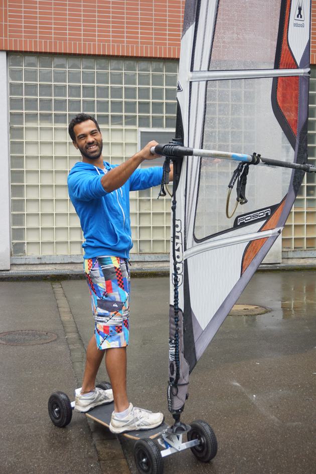 Tarek Sadek steht auf einem Skateboard mit Surfsegel. Er hält das Surfsegel und blickt lachend in Richung Kamera.