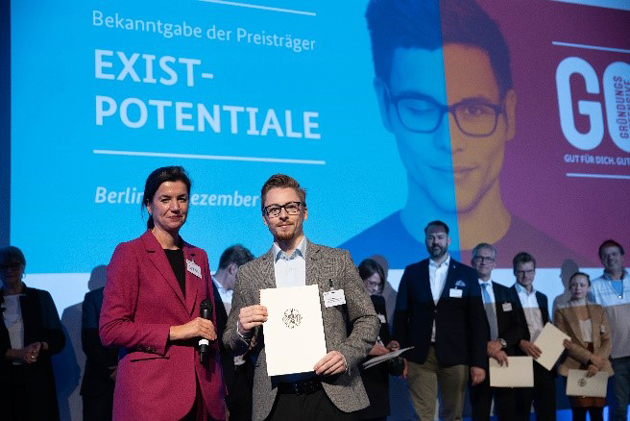 Sabine Hepperle, Abteilungsleiterin Mittelstandpolitik des Bundesministeriums für Wirtschaft und Energie, überreicht Felix Duffner, Mitarbeiter von Kilometer1, auf einer Bühne eine Urkunde. Im Hintergrund steht mit weißer Schrift auf blauem Hintergrund: "EXIST-POTENTIALE".