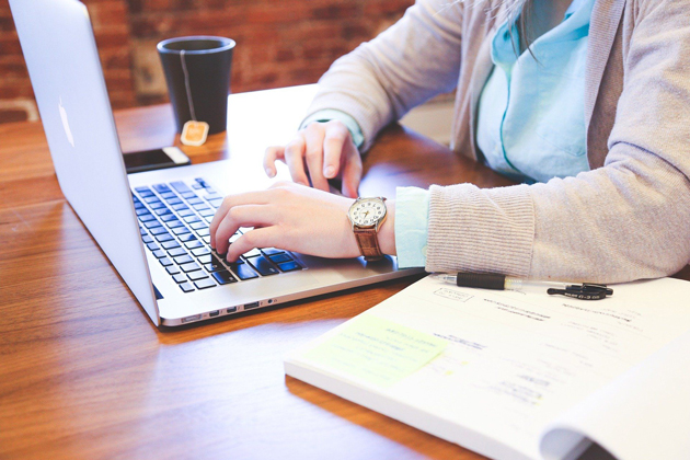 Zu sehen ist der Oberkörper einer Frau in einer blauen Bluse mit einer rosa Weste. Sie sitzt an einem Holztisch und arbeitet an einem aufgeklappten Laptop.