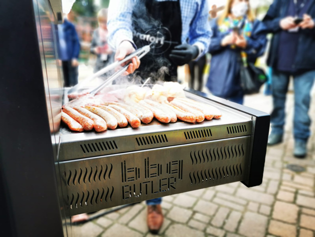 Blick von der rechten Seite auf eine ausgefahrene Grillplatte, auf der Würstchen liegen. Die Grillplatte sieht aus wie eine Art Schublade, die nach vorne aus einem Schrank, dem BBQ-Butler, ausgefahren ist.