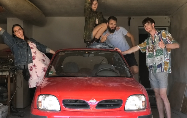 Blick in eine Garage, in der ein etwas älteres Modell eines roten Kleinwagens stet. Aus dem rechten Fenster beugt sich eine junge Frau. Auf dem Dach und links des Autos stehen bzw. sitzen drei junge Männer.