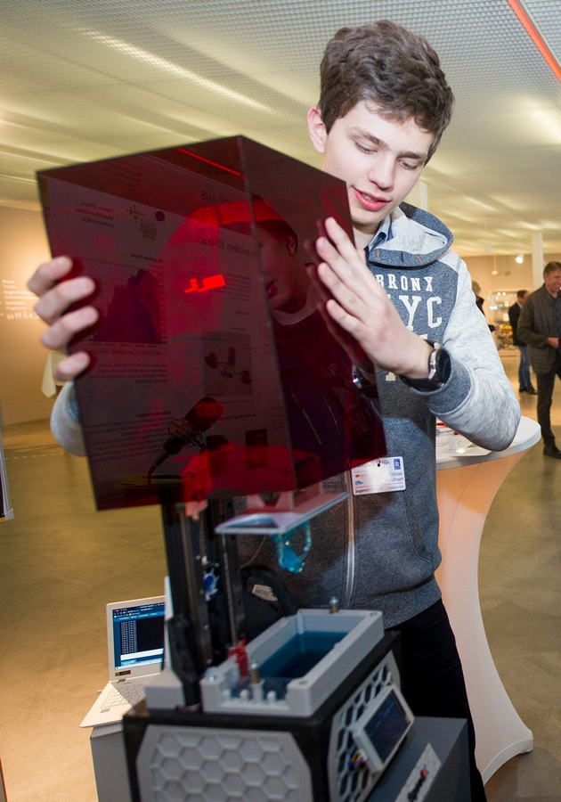 Ein junger Mann hebt einen roten Plexiglaskasten hoch. Darunter kommt ein 3D-Drucker zum Vorschein.