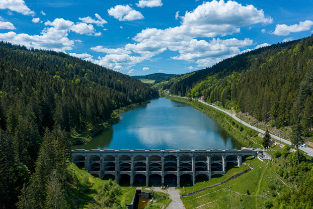 Luftaufnahme auf das Staubecken. Rechts und links des Wassers erheben sich bewaldete Berge, vorne ist die Staumauer zu sehen.