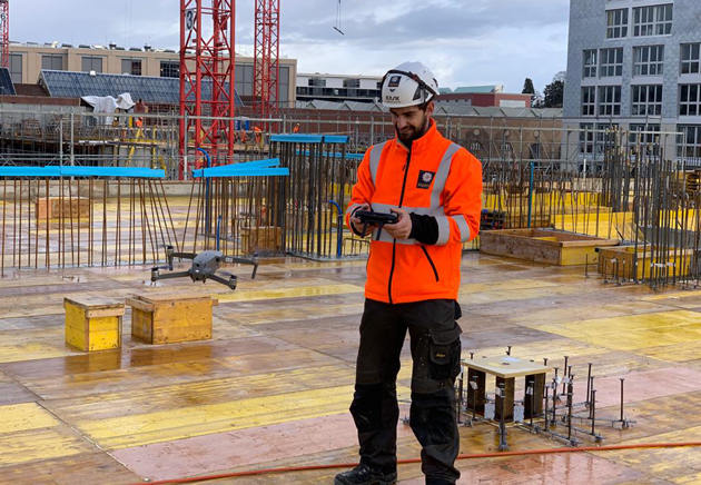 Sebastian Mattes steht auf einer Baustelle und steuert mit einer Fernsteuerung eine Drohne.