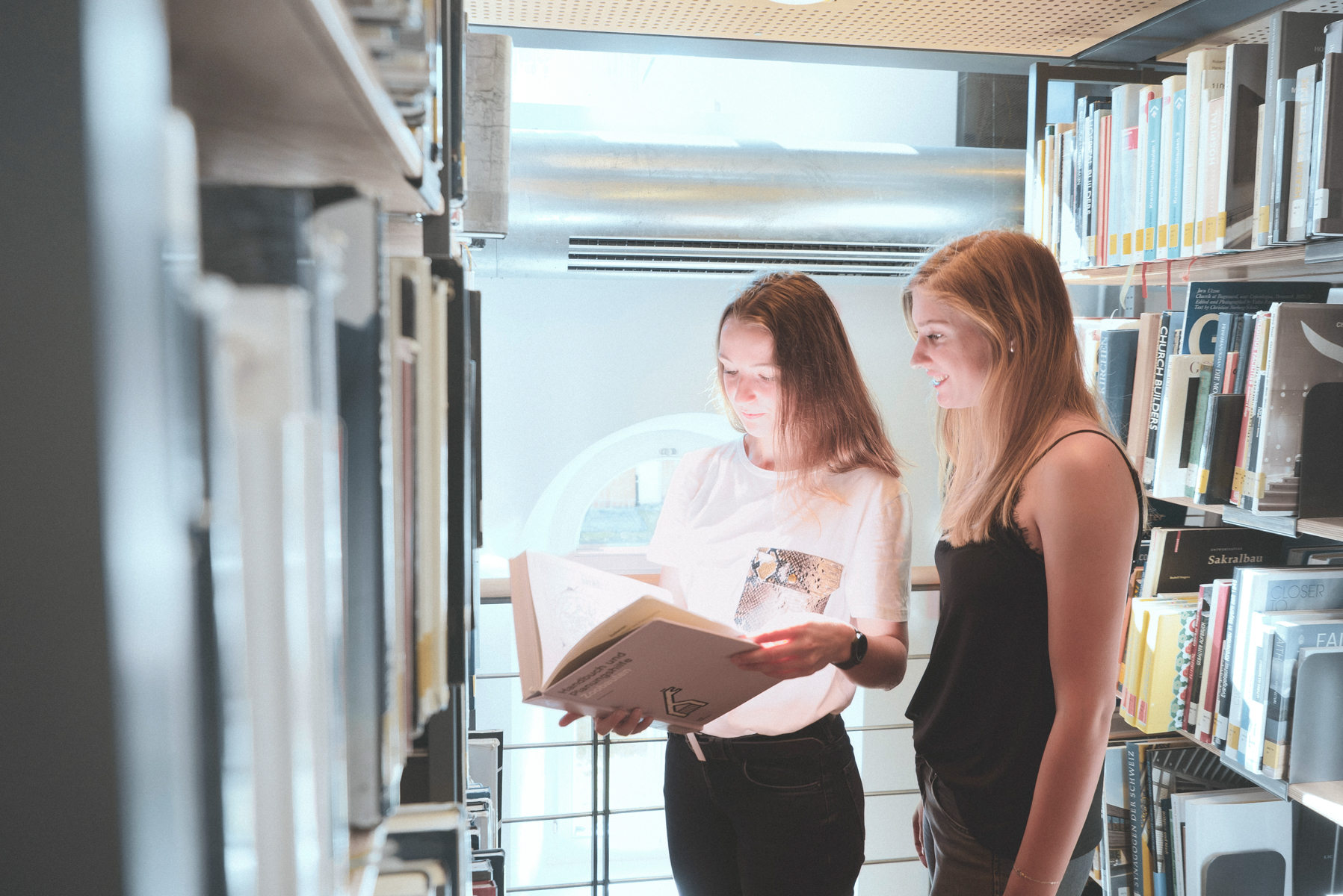 Zwei Studientinnen stehen zwischen Bücherregalen in der Bibliothek und schauen gemeinsam in ein großes Buch.