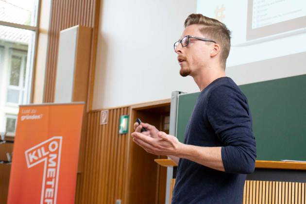 Felix Duffner arbeitet nach seinem Master-Studium an der HTWG nun in der Gründerinitiative Kilometer1. Daneben arbeitet er im Marketing für Unisphere. Vor einer grünen Tafel ist der Oberkörper von Felix Duffner von der Seite zu sehen. Links neben ihm steht ein Roll-up der Gründerinitiative Kilometer1.
