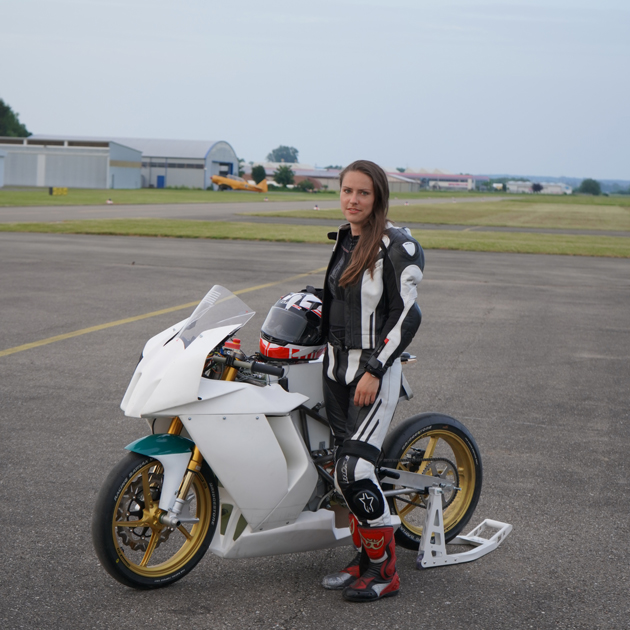 Eine junge Frau steht auf einem Flugfeld in Motorradkleidung neben einem Motorrad. Sie hält den Helm in der Hand. Im Hintergrund ist ein Hangar zu erkennen.