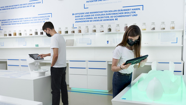 Eine Studentin und ein Student stehen in einem Ausstellungsraum, der mit kaltem Licht ausgeleuchtet ist. Sie blicken auf Tablets, die sie vor Ausstellungsstücke halten.