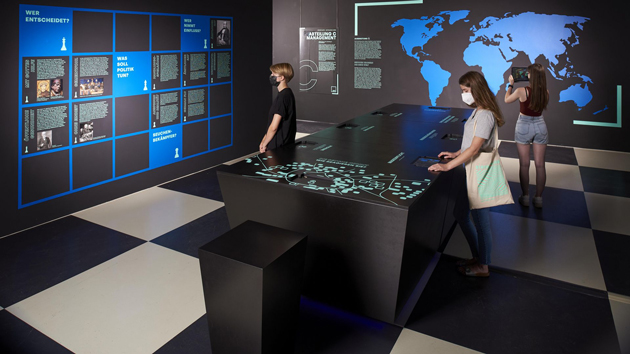Zwei Ausstellungsbesucherinnen stehen in einem Raum mit überwiegend schwarzen Wänden. Auf einer Wand ist eine blaue Weltkarte zu sehen.