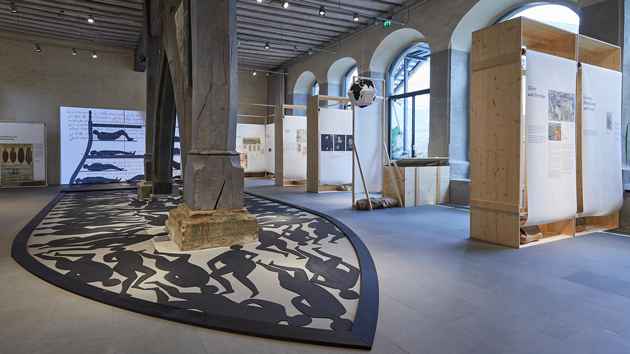 Blick in einen Ausstellungssaal mit einem alten Holzpfosten, um den sich auf dem Boden ein Schiffsrumpf mit den schwarzen Schatten von Menschen rankt.