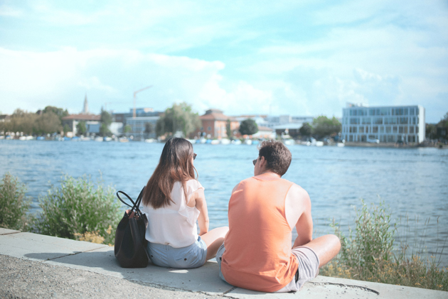 Blick auf die Rücken von einer jungen Frau und einem jungen Mann, die am Seerhein-Ufer sitzen. Im Hintergrund ist der Campus der HTWG zu sehen.