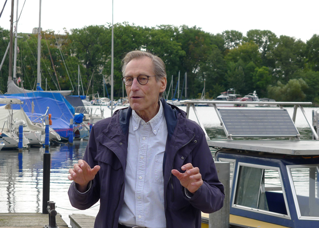 Prof. Christian Schaffrin steht mit den Händen gestikulierend am Hafen. Im Hintergrund ist das Boot Korona im Tegeler See zu sehen.
