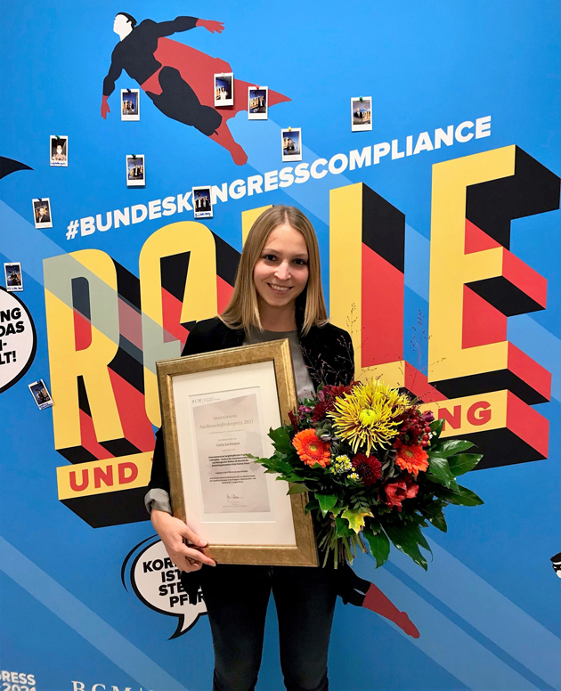 Eine junge Frau steht vor einem tiefblauen Banner. Sie hälte eine eingerahmte Urkunde und einen Blumenstrauß in ihren Händen.