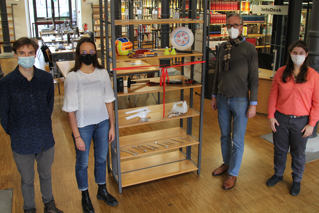 Zwei Studentinnen und ein Student stehen mit dem Leiter der Bibliothek neben einem Regal in der Bibliothek der HTWG. Im Regal liegen Küchen- und Sportgeräte sowie Werkzeug.