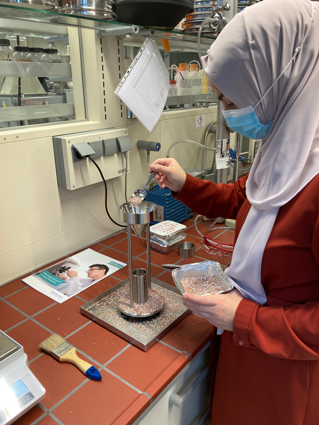 Eine junge Frau steht an einem Labortisch. Sie gibt Materialaufschluss zur Schüttdichtebestimmung in ein Gerät.