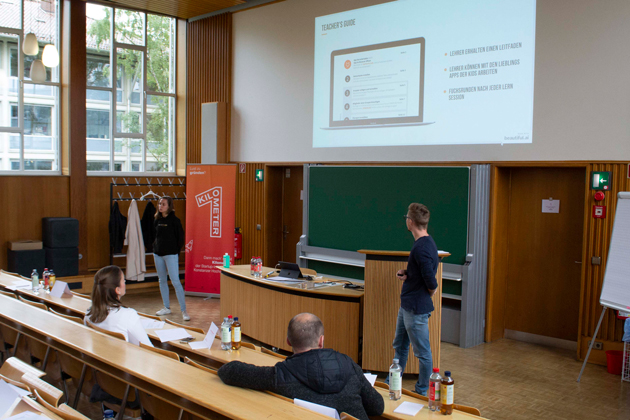 Eine junge Frau und ein junger Mann stehen in einem Hörsaal vor dem Auditorium und erläutern eine Präsentation.