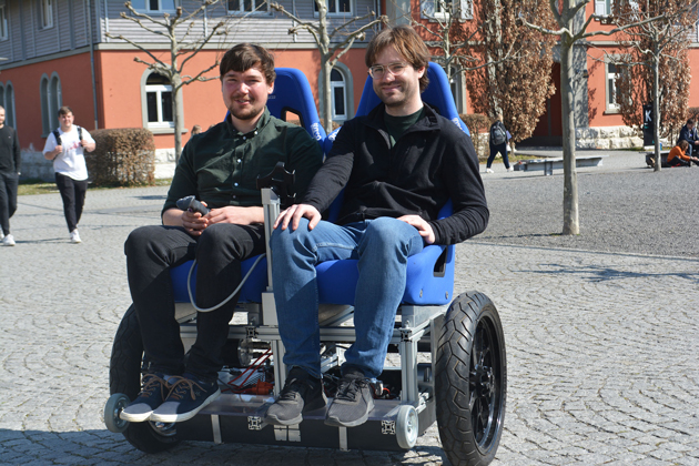 Ein doppelsitziges Zweirad fährt über den Campus der HTWG. In den Sesseln des Gefährts sitzen links Hannes Homburger, rechts Patrick Hoher.