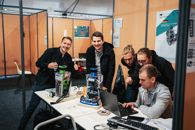 Eine Gruppe junger Männer steht um einen Tisch. Auf dem Tisch stehen ein Roboter und ein Laptop.