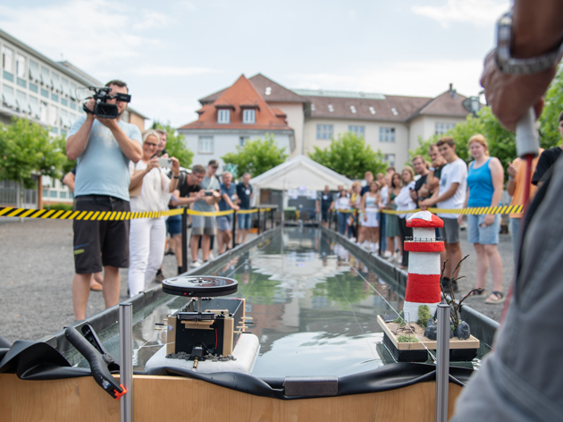 Blick auf eine 16 Meter lange Wasserwanne, in der zwei Solarboote liegen. Rechts und links schauen Menschen auf die Boote.