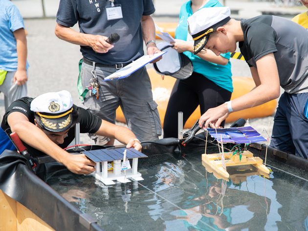 Schülerinnen mit Kapitänsmütze beugen sich über ihre Modellboote, die im Wasser schwimmen.