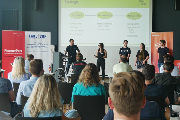 Eine Gruppe von fünf Studierenden präsentiert in einem Saal ihr Gründungskonzept. Aufmerksame Hörder*innen sind von hinten zu sehen.