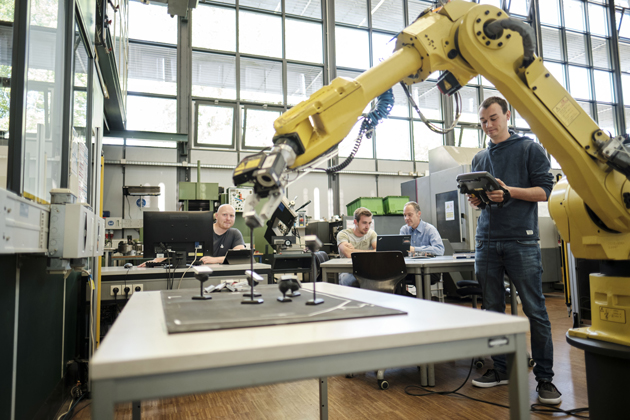 Im Vordergrund ist ein großer Roboterarm zu sehen. Dahinter sitzen Studierende an Tischen und arbeiten an ihren Laptops mit Blick auf den Roboterarm.