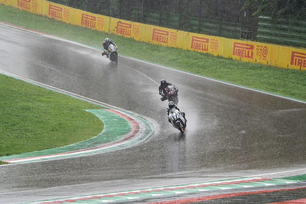 Drei Motorräder liegen während eines Rennens auf regennasser Fahrbahn in einer Kurve. 