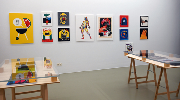 Blick in einen Ausstellungsraum mit Illustrationen an der Wand und zwei Ausstellungstischen.