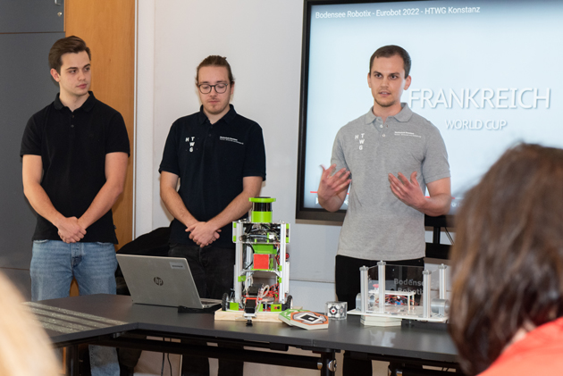 Drei junge Männer stehen zwischen einer Präsentation auf einer Leinwand und einem Tisch. Auf dem Tisch steht ein Roboter.