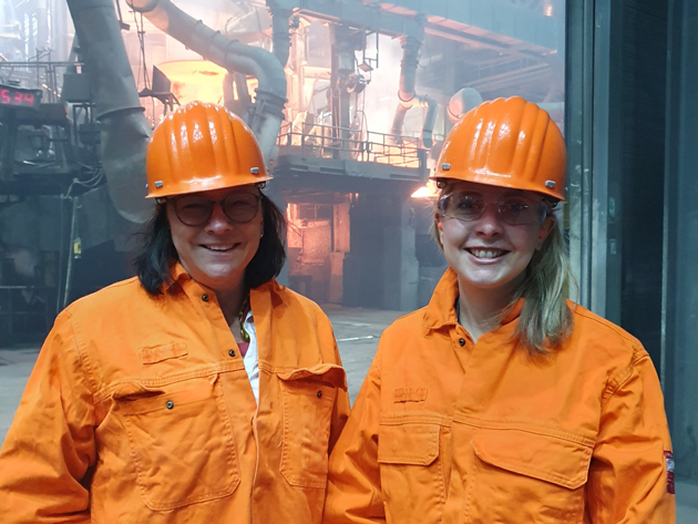 Zwei Frauen stehen in orangener Jacke und mit orangenem Helm in einer Industrieanlage vor einem Kupolofen.
