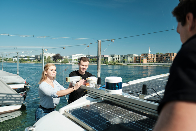 Eine junge Frau und ein junger Mann stehen an einem Boot. Im Hintergrund ist der Seerhein zu sehen. Auf dem Boot sind Solar-Paneelen und ein Lidar-Sensor zu sehen.