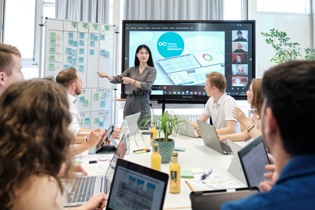 Szene eines Meetings. Eine Frau präsentiert vorn an einem Bildschirm. Eine Gruppe junger Leute mit Laptop vor sich auf dem Tisch blickt auf sie.