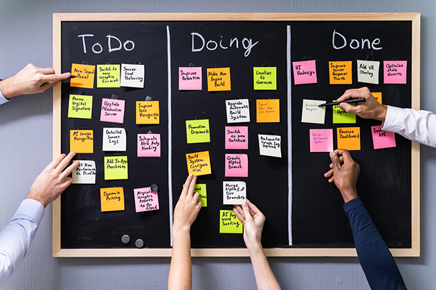 Das Bild zeigt eine schwarze Tafel, die in die Arbeitsbereiche To Do, Doing und Done aufgeteilt ist. In jedem Arbeitsbereich kleben bunte Post-Its, auf denen Aufgaben beschrieben sind. Hände von drei Personen fassen Post-Its an und verschieben sie auf der Tafel.