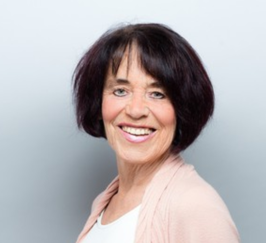 Annette_Kleinfeld_2019 Sie trägt einen kurzen, schwarzen Bob, ein weißes Oberteil mit einer rosa Jacke und blickt nach vorne.