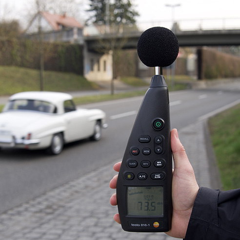 Blick auf eine Straße. Auf der Fahrbahn ein weißes Auto, in der rechten Bildhälfte streckt eine Hand ein Messgerät in Richtung Fahrbahn.