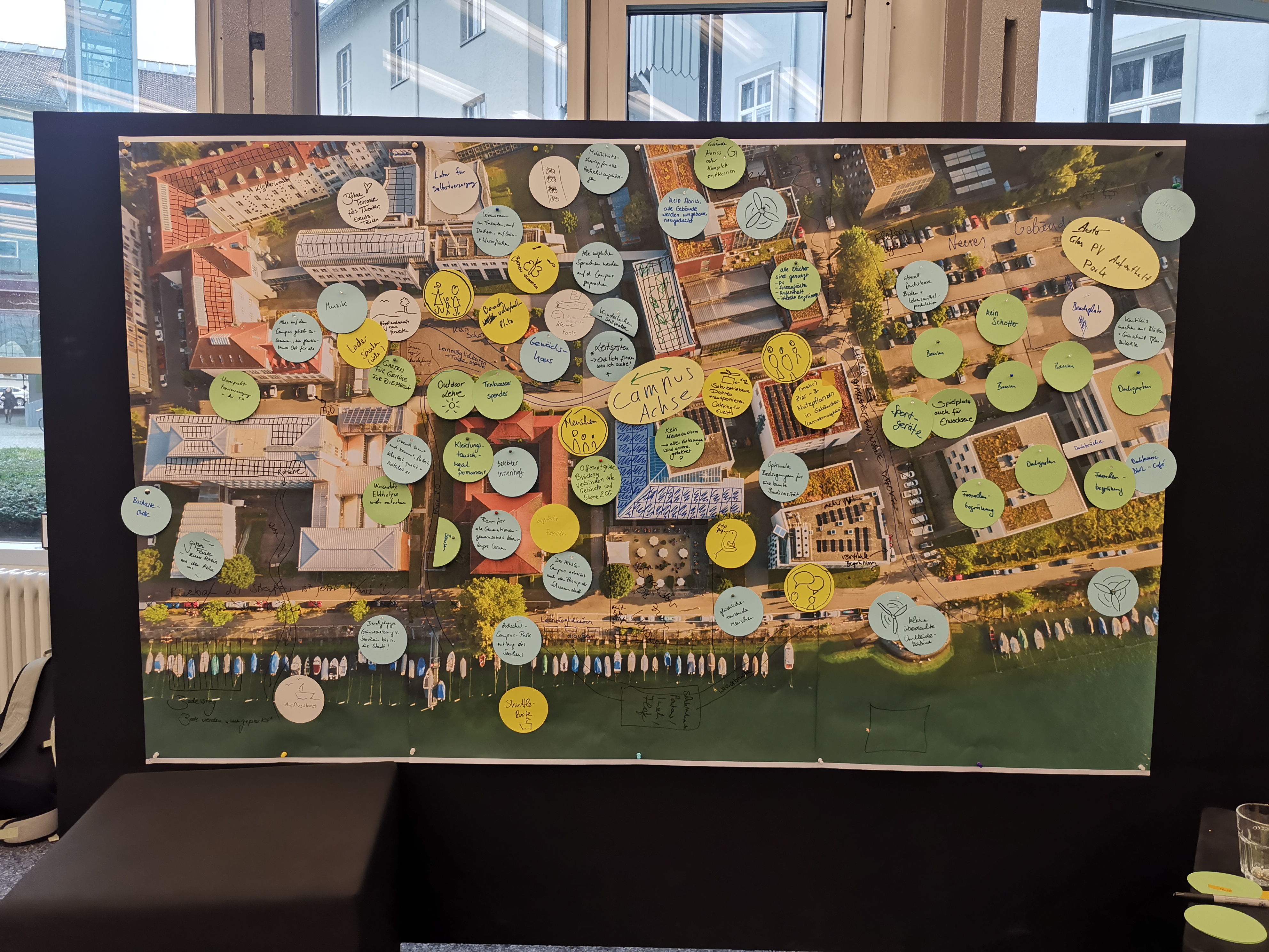 Das Bild zeigt ein großes Plakat einer Luftaufnahme des Campusgeländes, auf welches die Ideen zum klimapositiven Campus gezeichnet und geklebt wurden. In einem Workshop zum ZielBILD der klimapositiven HTWG 2030 wurde eine Utopie der HTWG entworfen. Die Ideen werden in eine interaktive Campuskarte eingebettet.