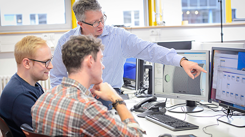 A professor explains something to two students on a computer monitor