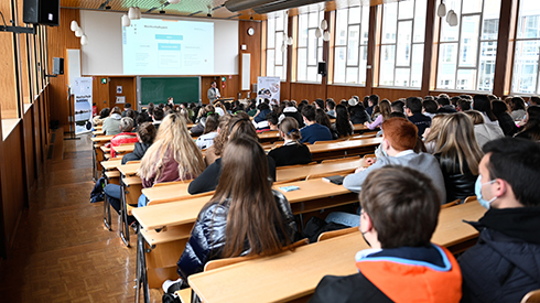 Schüler*innen lauschen in einem Vorlesungssaal dem Vortrag eines Professors (von hinten fotografiert).